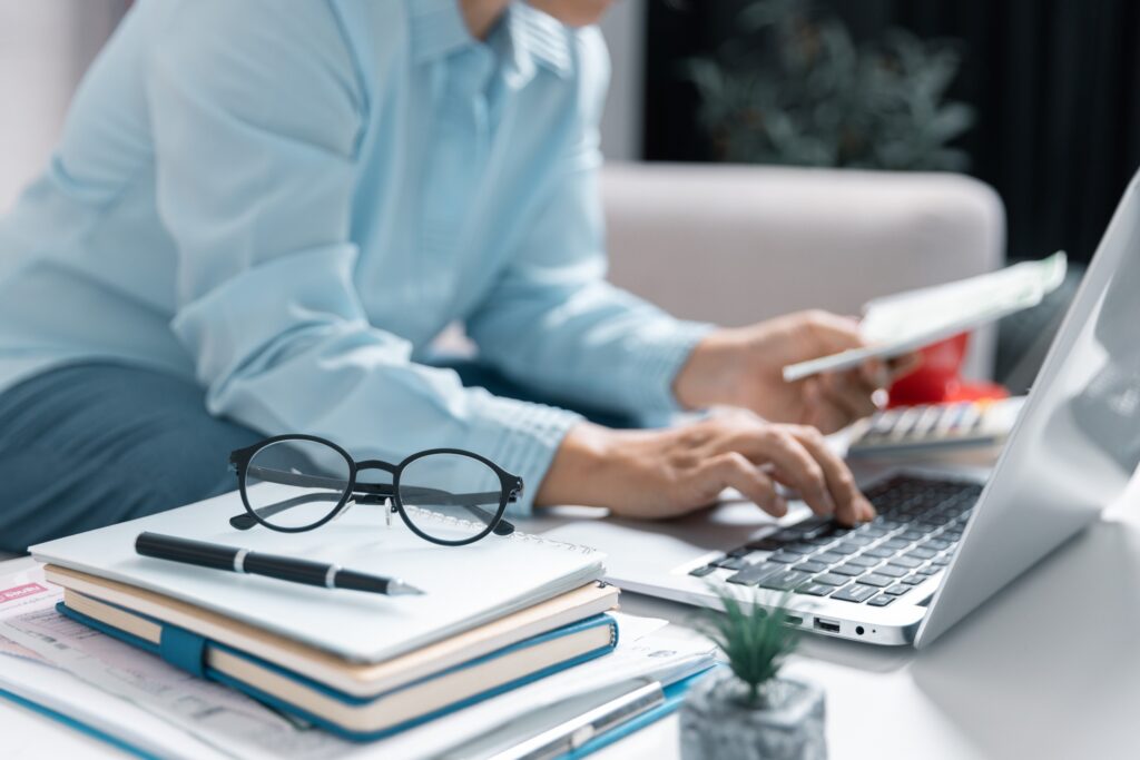 A man researching how to become HR certified on his laptop next to a stack of notes, glasses, and a pen