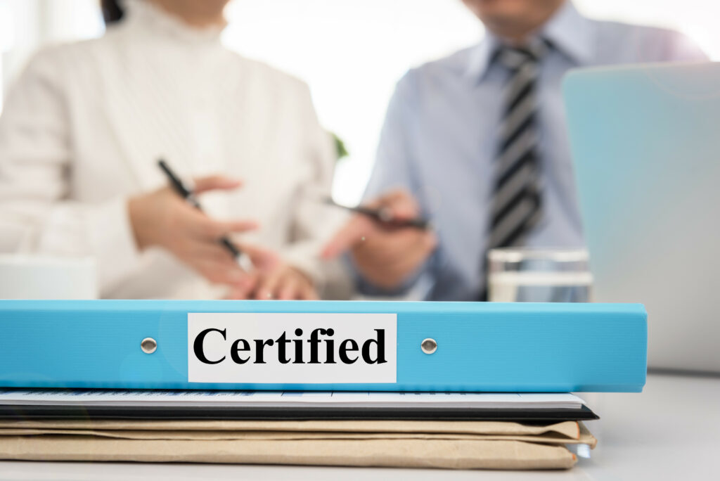 Blue binder labeled 'Certified' on a stack of documents with two business professionals discussing paperwork in the background